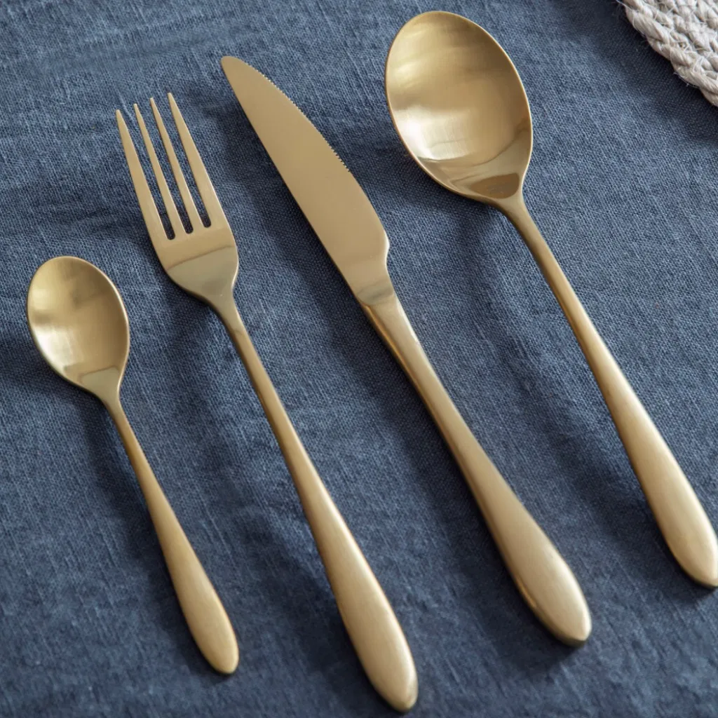 Four pieces of contemporary 16-piece cutlery set in a smooth, matte brass or brushed gold finish, including a fork, knife, dinner spoon, and teaspoon, resting on a dark blue linen tablecloth.