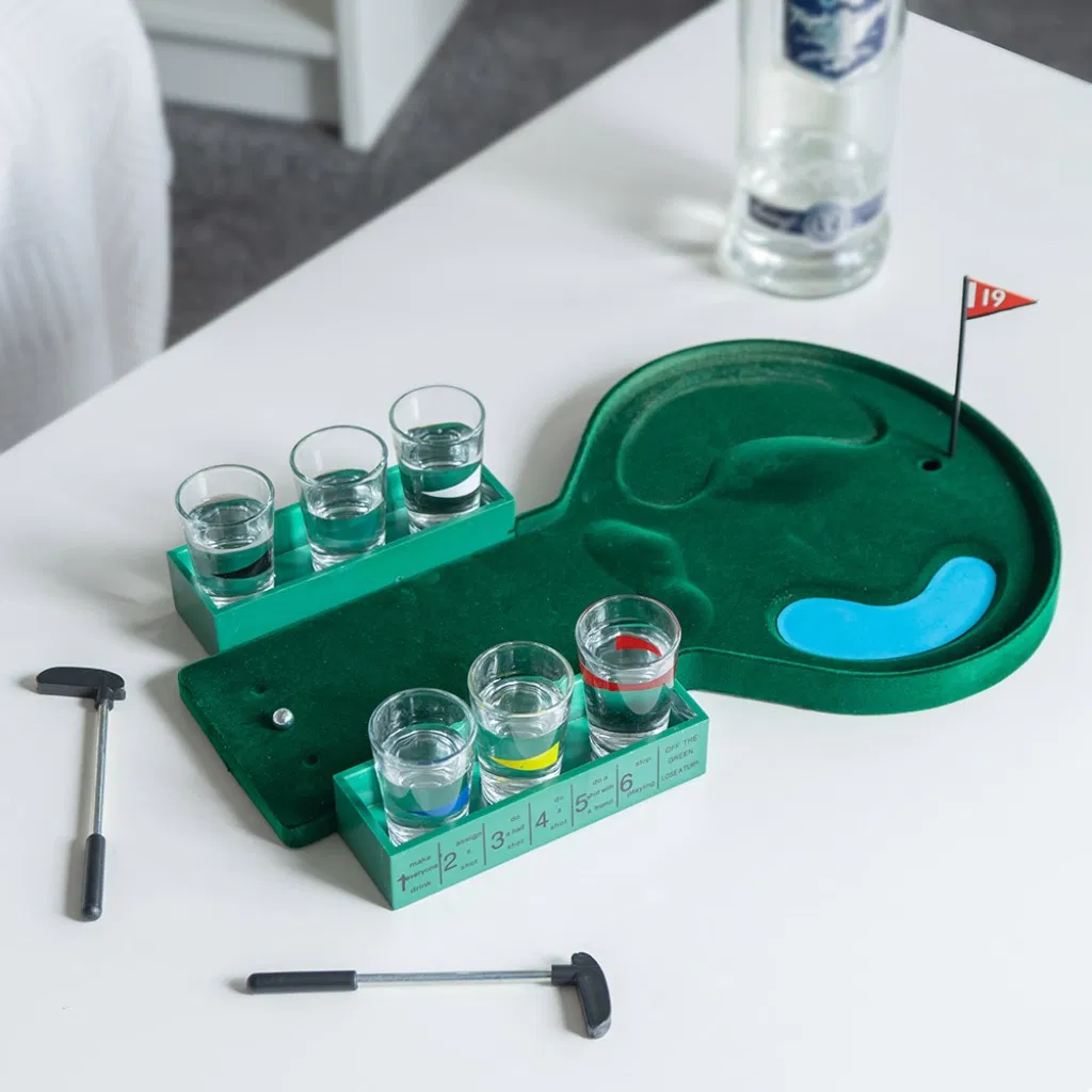 19th Hole miniature golf drinking game set, featuring a green felt putting surface, six shot glasses, two miniature putters, and a flag, displayed on a white table.