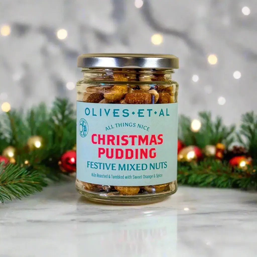Glass jar of Christmas Pudding Festive Mixed Nuts, Kiln Roasted and tumbled with sweet orange and spice, set against a background of festive greenery and blurred bokeh lights.