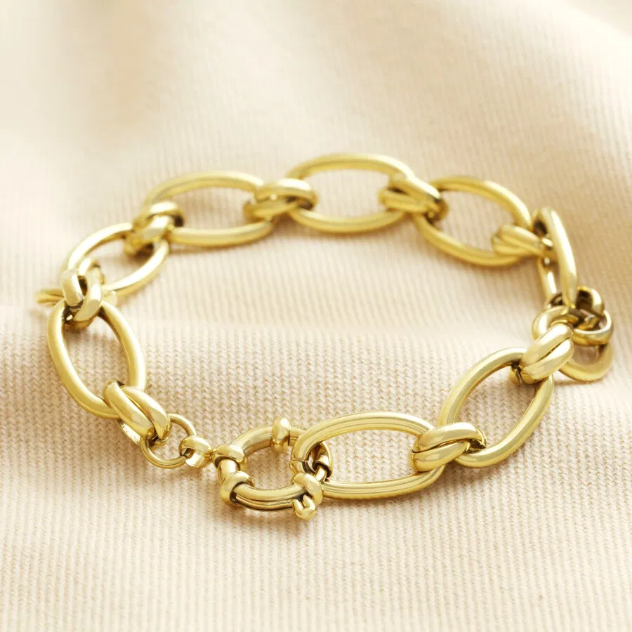 A gold-toned, chunky chain link bracelet made from large, interconnected oval links and secured with a lobster clasp, displayed against a wrinkled pale beige fabric background.