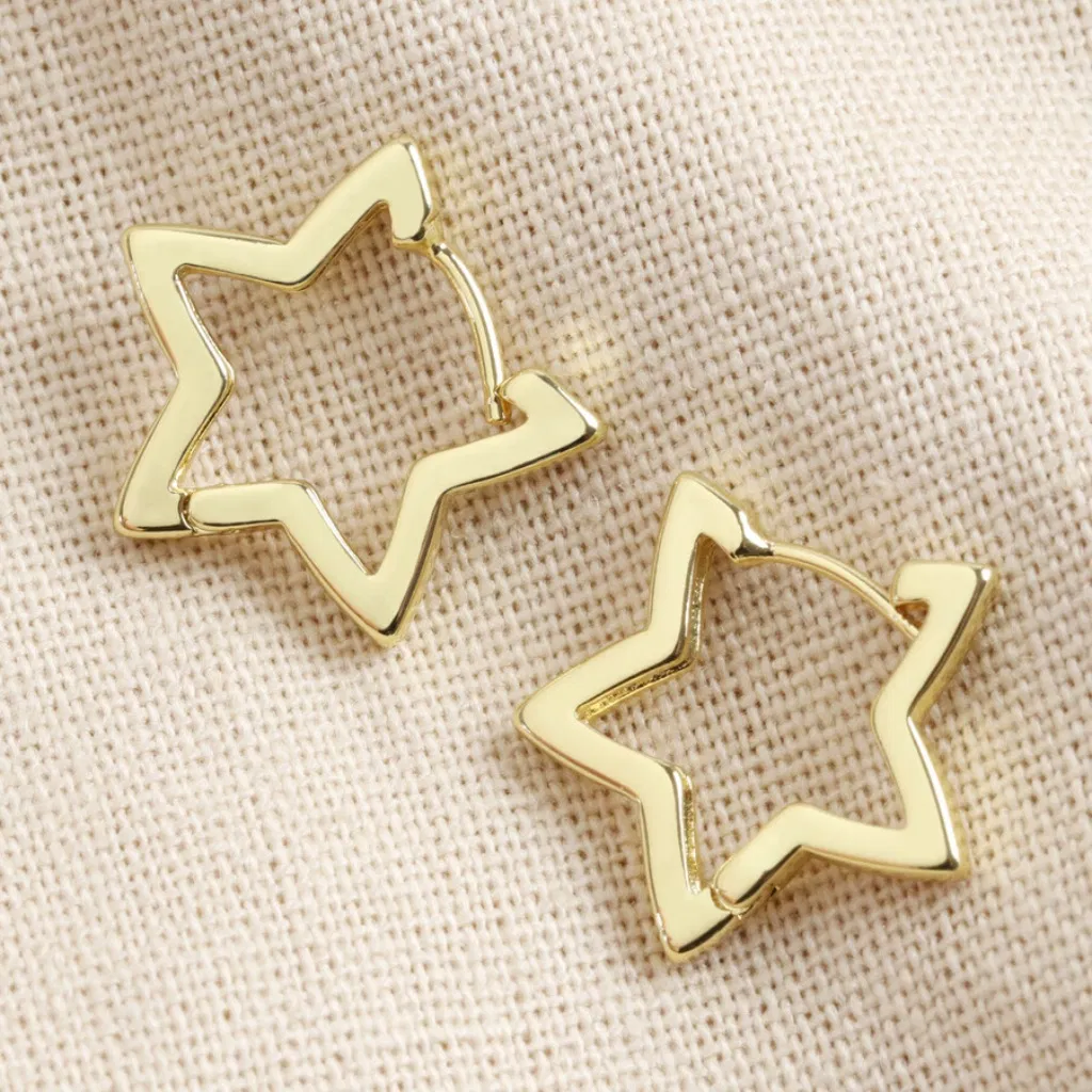 Close-up of a pair of gold-plated open star-shaped hoop earrings with a polished finish, resting on a neutral, textured beige fabric, highlighting the modern, celestial design.