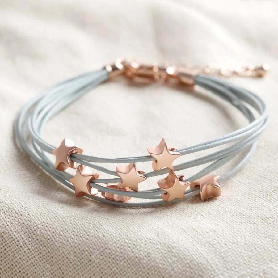 A delicate multi-strand bracelet featuring thin, grey faux leather cords adorned with several small, three-dimensional rose gold-toned star beads, secured with a rose gold lobster clasp and displayed on a textured beige fabric background.
