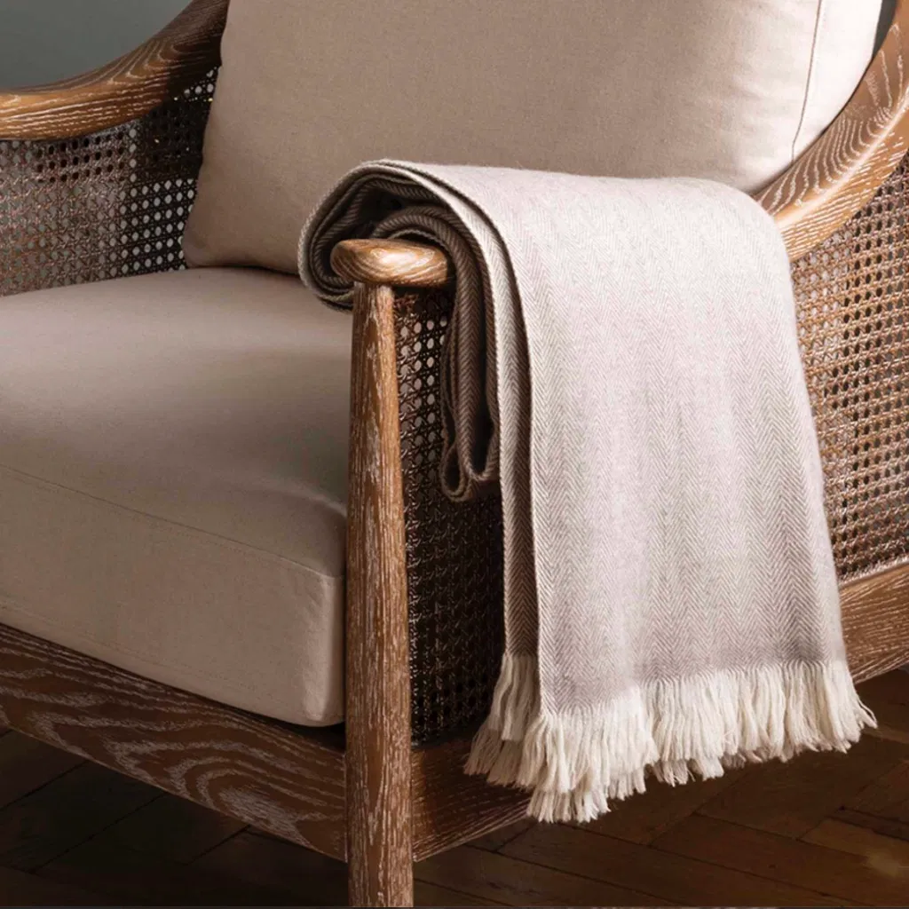 Luxury 100% New Zealand wool throw blanket in an oatmeal or pale sage green herringbone pattern with a thick cream fringe, folded and draped over the arm of a wooden armchair with cane webbing.