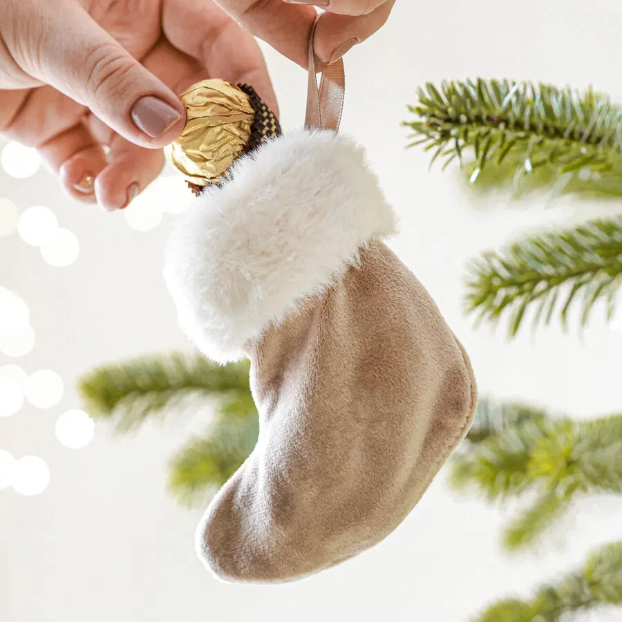 A small, beige velvet Christmas stocking hanging ornament with a white faux fur cuff and a brown ribbon loop, being held by a hand placing a gold-wrapped chocolate into the opening, set against a blurred background of a Christmas tree and fairy lights.
