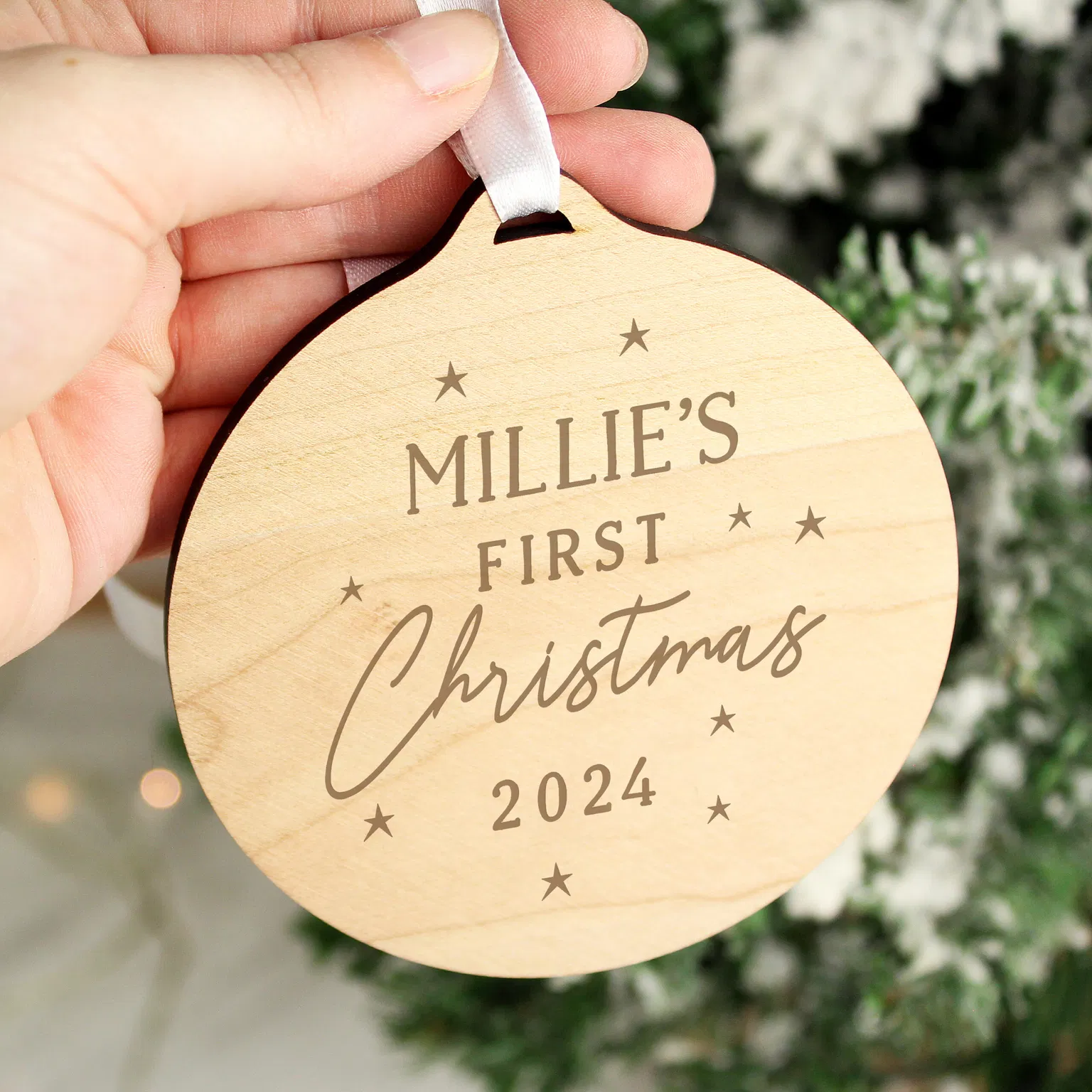 Hand holding a round, wooden "Baby's First Christmas" tree decoration, personalized with the name Millie's and the year 2024, engraved with small stars, hanging near a flocked Christmas tree.