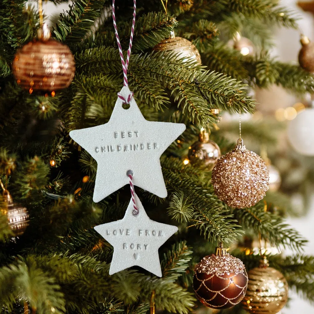 Two personalised handmade clay or ceramic star-shaped Christmas tree decorations, one engraved 'BEST CHILDMINDER' and the smaller one 'LOVE FROM RORY', hanging from a red and white string on a lush green Christmas tree adorned with copper and gold baubles.