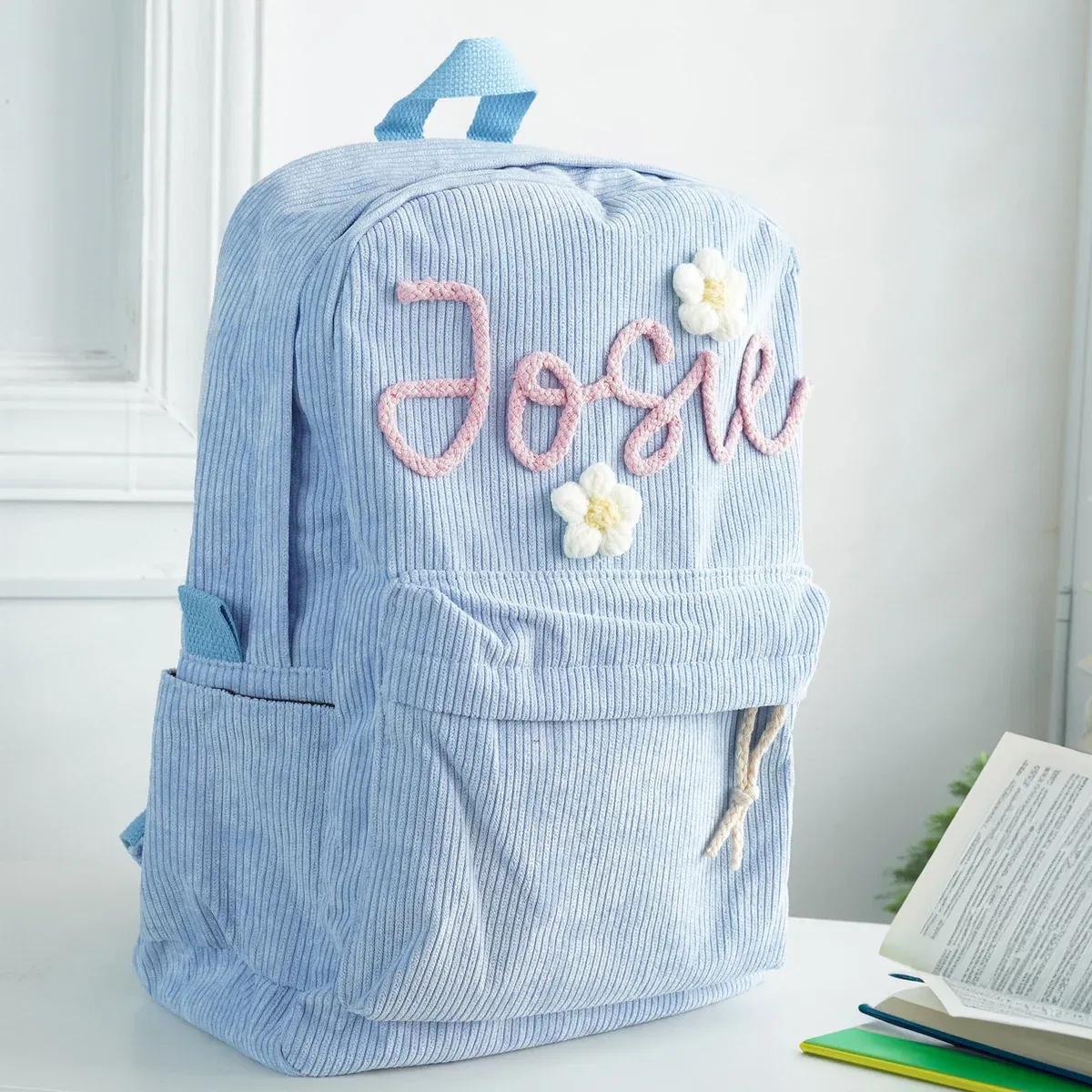A light blue corduroy children's backpack shown indoors. The bag is personalised with a large, pink script embroidery of the name "Josie" on the front, accented by small white embroidered flowers. The backpack has a front pouch and side pockets.