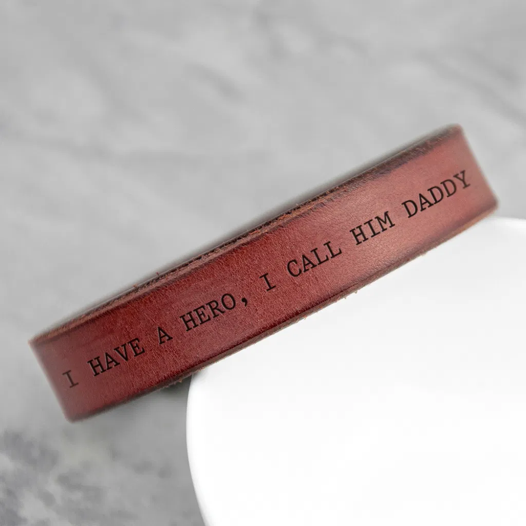 Close-up of a thick, personalized brown leather cuff bracelet with a custom engraved message, 'I HAVE A HERO, I CALL HIM DADDY', resting on a white surface.