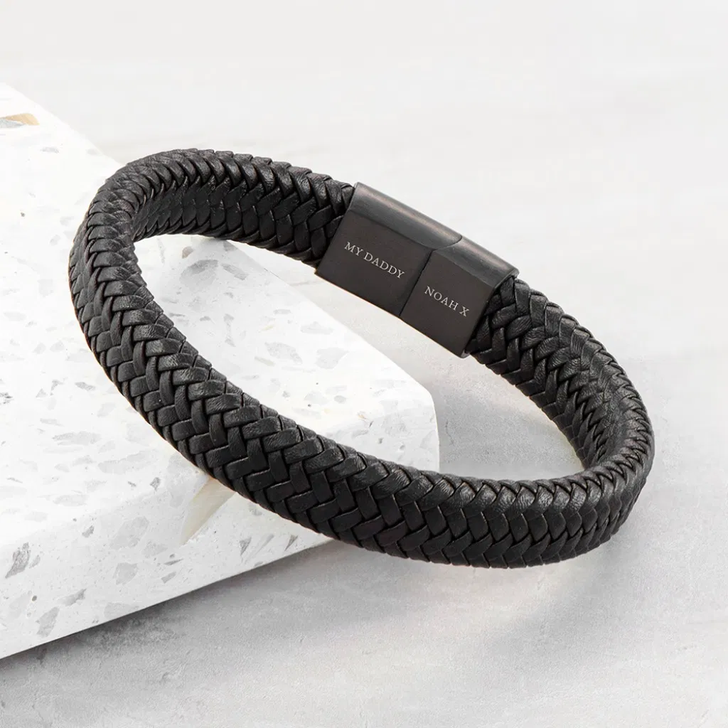 Close-up of a personalised men's black leather braided bracelet featuring a matte black magnetic clasp engraved with custom white text, such as 'MY DADDY' and 'NOAH X'.