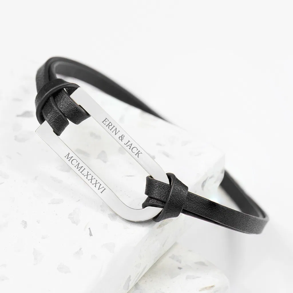 Close-up photograph of a personalised men's silver bar leather bracelet. The bracelet features a polished silver bar, engraved on the visible side with "ERIN & JACK" on the top line and the Roman numerals "MCMLXXXVI" on the bottom line. The silver bar is threaded through a thin black leather strap, which is tied in knots at both ends of the bar. The bracelet is displayed on a white, speckled surface.