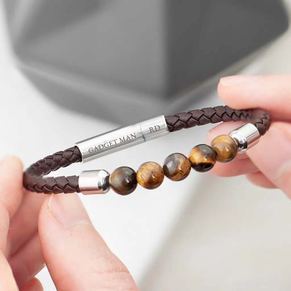 Close-up of a personalised men's bracelet featuring a brown woven leather strap, several polished Tiger's Eye gemstone beads, and a stainless steel clasp engraved with custom text like 'GADGET MAN'.