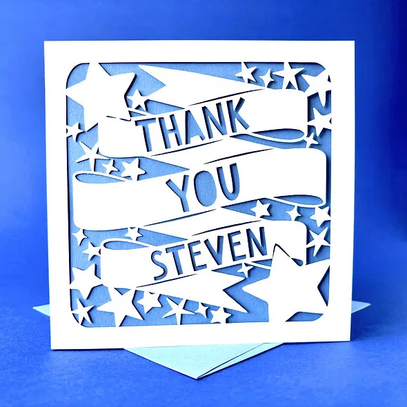 Close-up of a personalised thank you card featuring white laser-cut paper with large banners reading 'Thank You' and a custom name (Steven), surrounded by scattered stars, backed by a light blue insert.