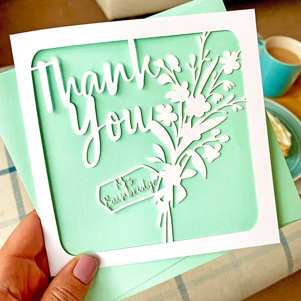 Hand holding a square, personalised thank you card featuring white laser-cut paper with a floral bouquet design and 'Thank You' script, backed by a mint green insert.