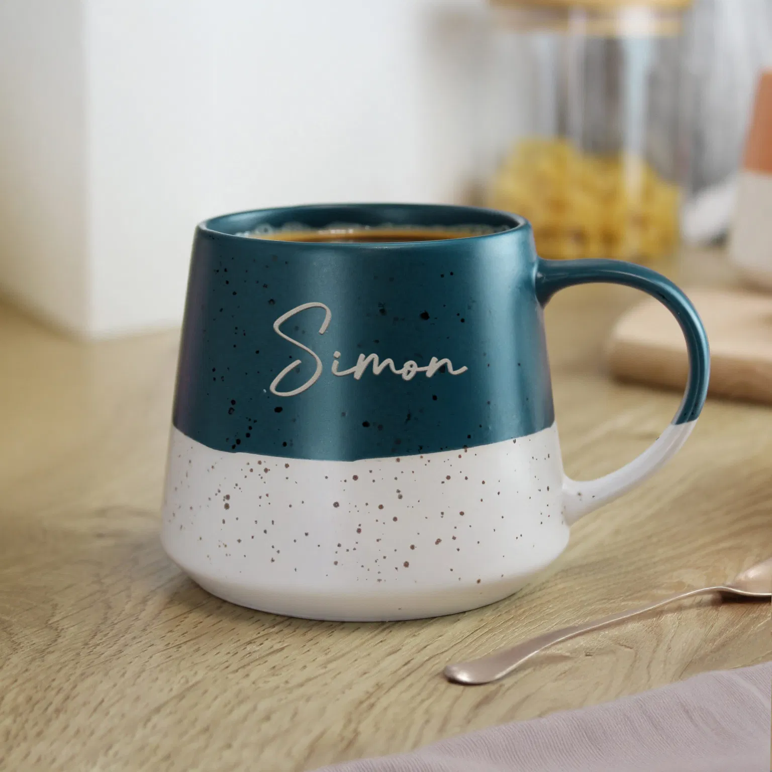Handcrafted stoneware coffee mug with a two-tone design: deep teal blue upper half and speckled white base, engraved with the custom name 'Simon' in a script font, displayed on a marble kitchen worktop.