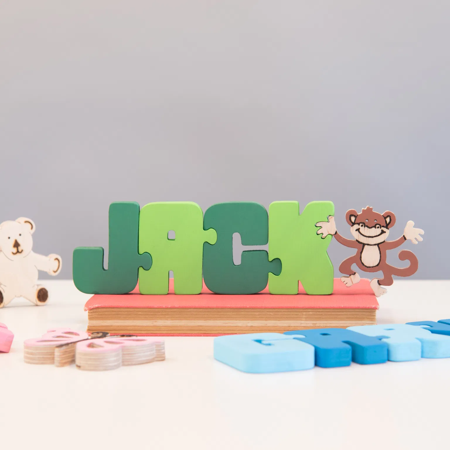 A colourful wooden name puzzle spelling the name 'JACK' in interlocking green letter pieces. The jigsaw stands on a pink book and is accompanied by small wooden decorative pieces, including a white bear and a brown monkey. This toy emphasizes personalized learning and bright, durable design. Ideal personalized gift for a toddler's birthday, Christening, or nursery decor.