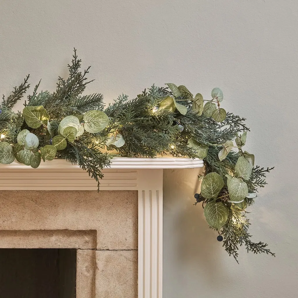 A close-up view of a Pre-Lit Christmas Garland featuring mixed faux evergreen, silvery-green eucalyptus, and dark berries, displayed on a classic white mantelpiece. Small warm white LED lights are glowing softly within the foliage, creating a cozy festive atmosphere.