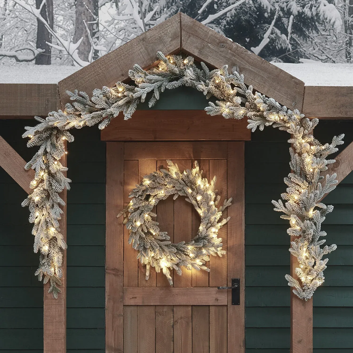 Pre-lit outdoor Christmas garland and matching wreath set, featuring snowy, flocked artificial pine branches with warm white lights, decorating a wooden door archway against a dark green shed and a snowy forest background.