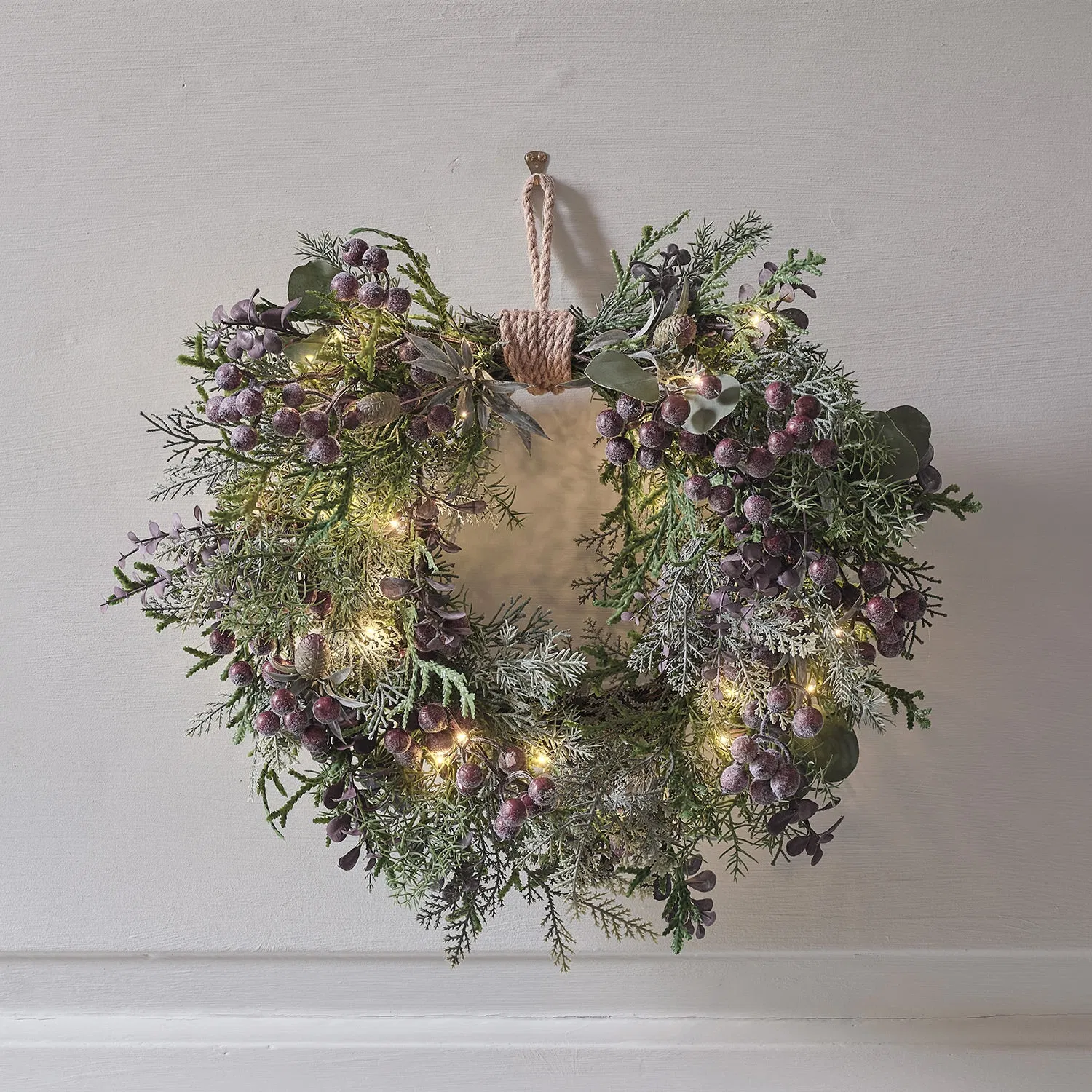 Decorative heart-shaped Christmas wreath featuring frosted purple berries, mixed artificial pine and cedar-like greenery, and warm white micro fairy lights, tied with jute rope and hanging on a pale wall.