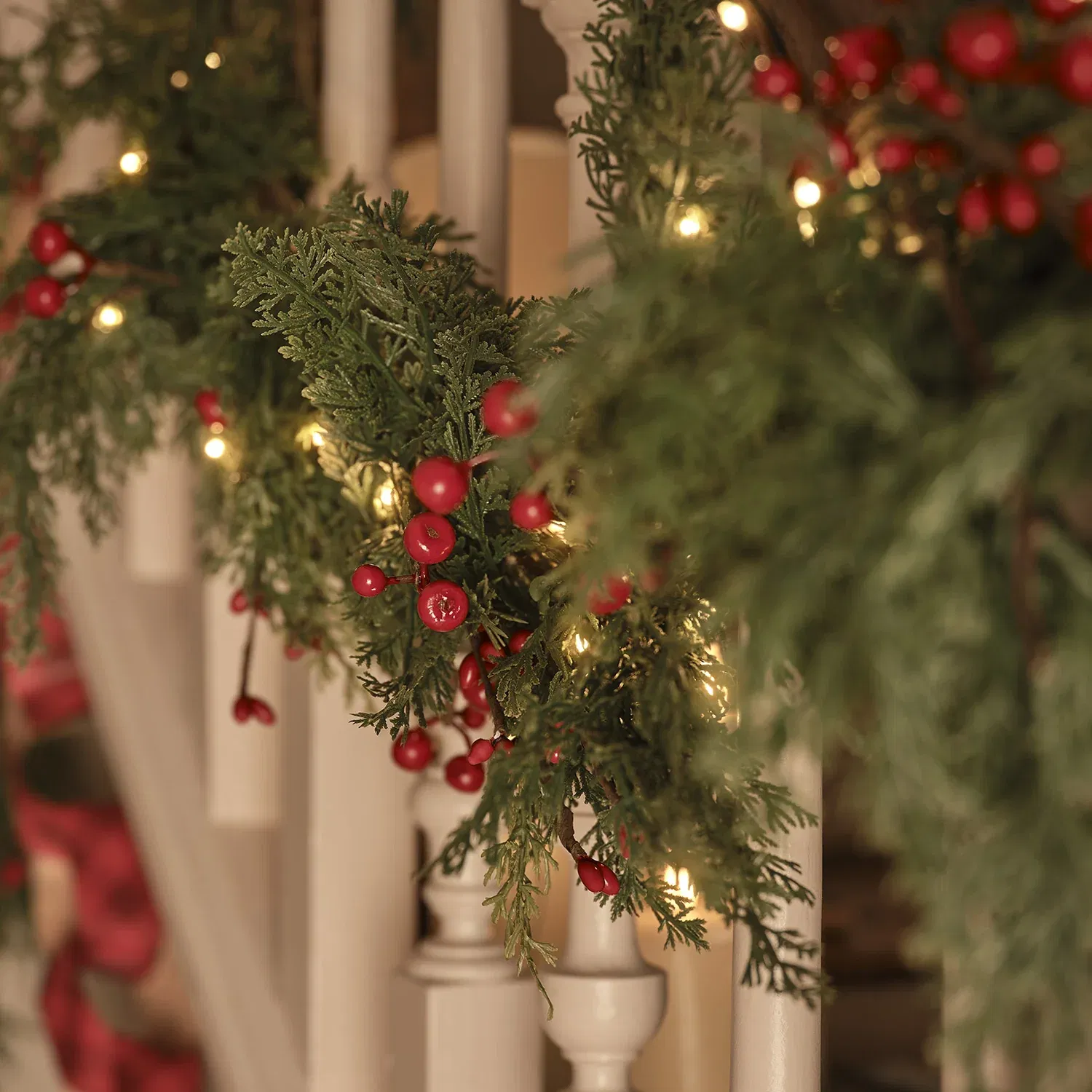 A close-up of a festive artificial Christmas garland featuring mixed green pine and cedar-like foliage, bright red berries, and integrated warm white micro fairy lights, resting horizontally on a light wooden countertop.