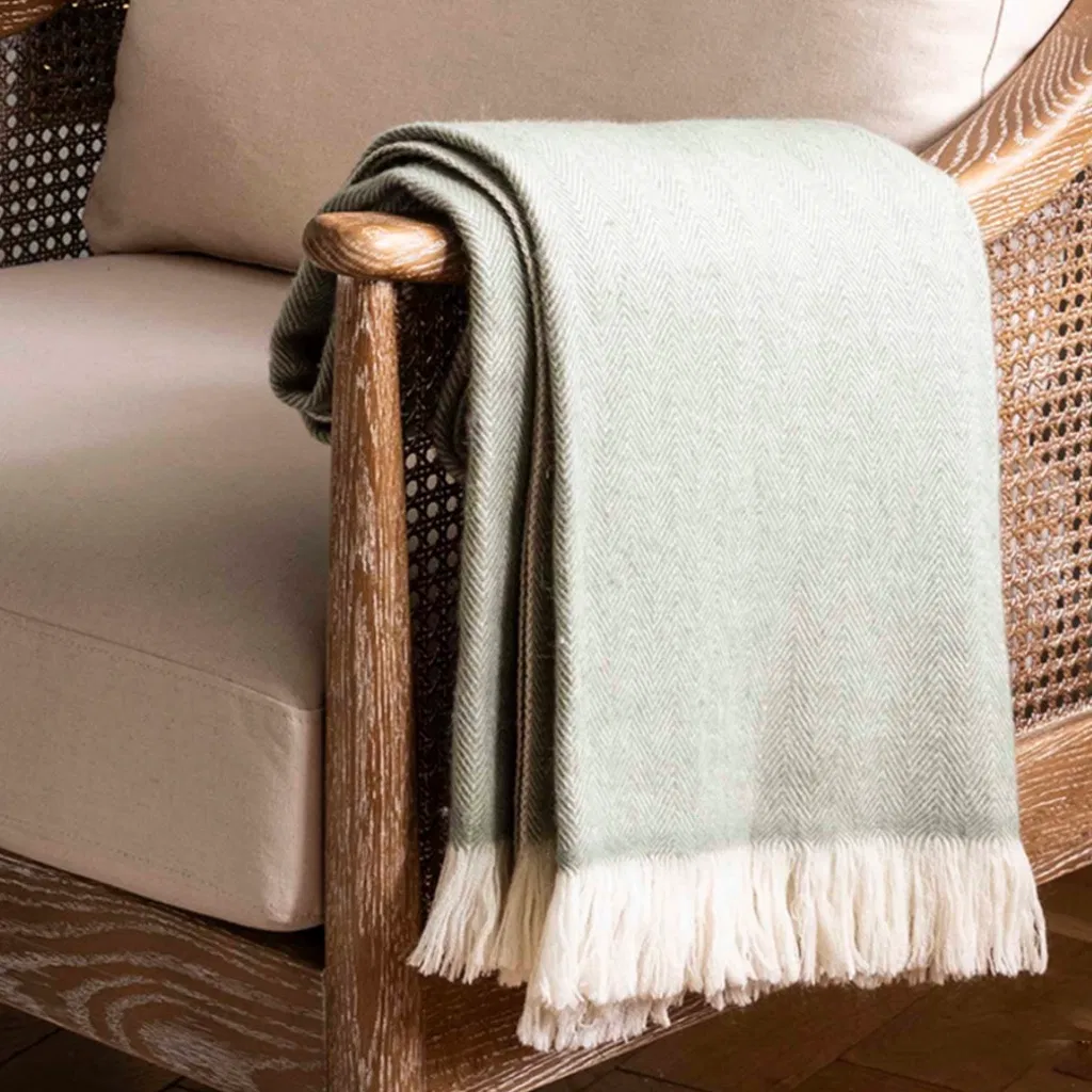 Luxury wool throw blanket in a pale sage green herringbone pattern with a thick cream fringe, folded and draped over the arm of a wooden armchair with cane webbing, creating a cosy Scandi look.
