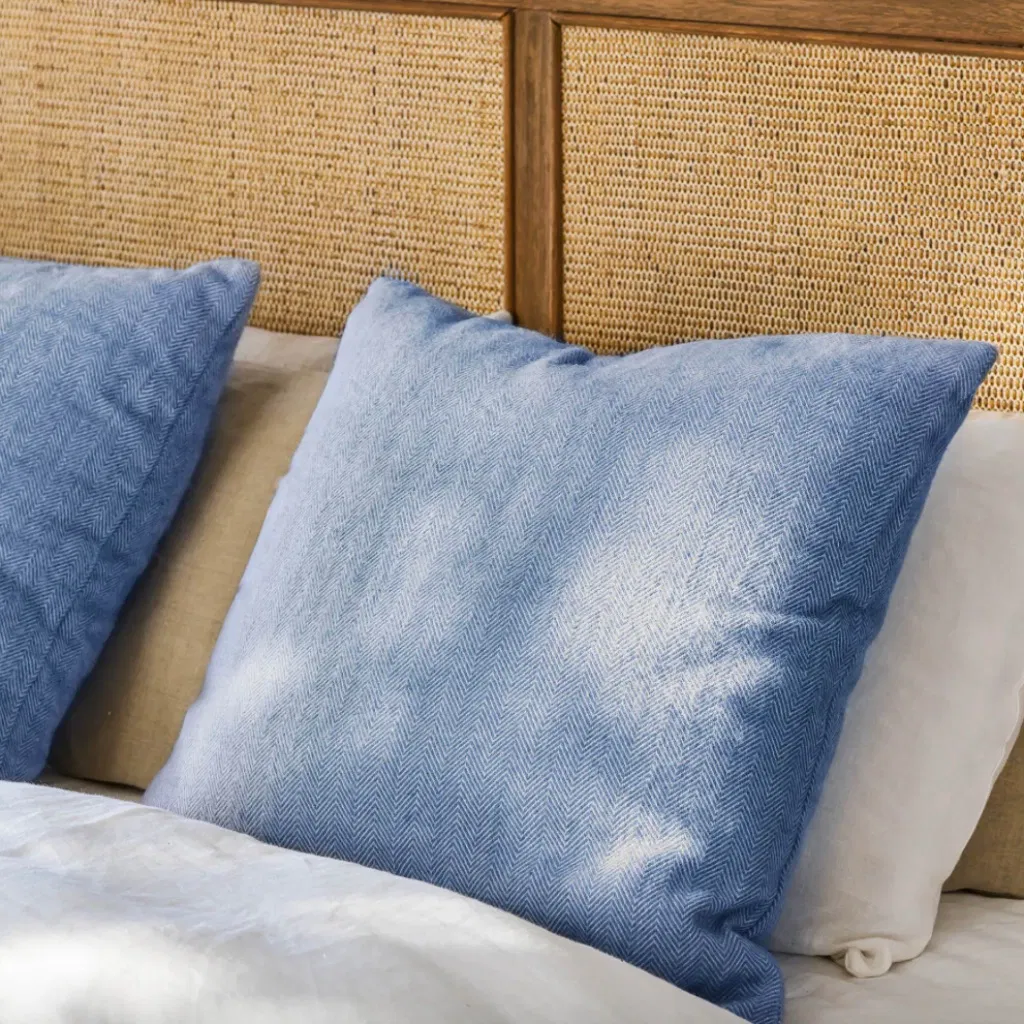 Square accent cushion in a cove blue or denim blue color with a visible subtle herringbone woven pattern, resting on a bed with a rattan cane headboard.