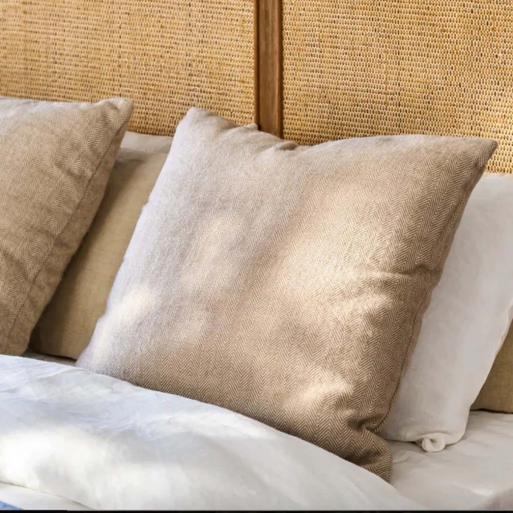 Square accent cushion in a natural oatmeal or light beige color with a textured herringbone woven pattern, resting on white bedding against a rattan cane headboard.