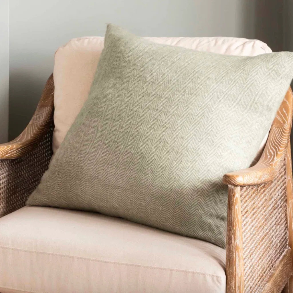Square accent cushion in a muted sage green color with a visible subtle herringbone woven pattern, resting on a beige armchair with a natural cane frame.
