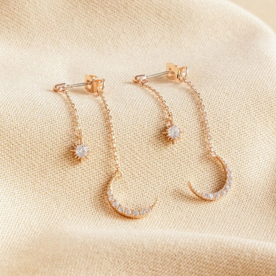 A pair of rose gold-toned double drop earrings, featuring a small crystal star charm connected by a delicate chain to a larger crystal-studded crescent moon charm, displayed on a pale, textured cream fabric background.