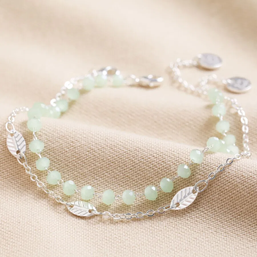 A set of two delicate silver bracelets displayed together, one featuring a continuous chain of pale mint green crystal beads, and the other a fine silver chain linked with multiple small, textured silver leaf charms. Both bracelets have a small round charm at the clasp.