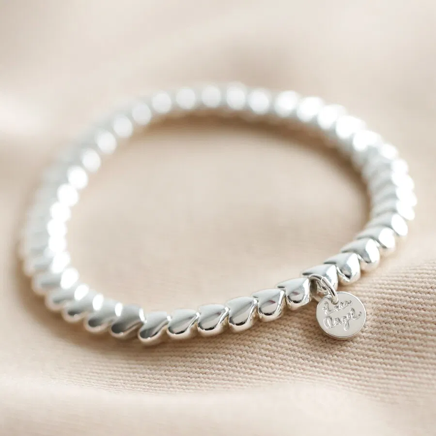 A delicate silver stretch bracelet composed entirely of interlocking, smooth silver heart-shaped beads, featuring a small, round silver charm with a subtle inscription hanging near the clasp, displayed on a textured pale fabric background.