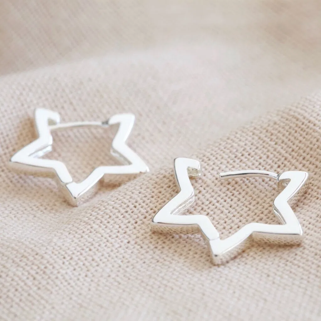 Close-up of a pair of delicate silver open star-shaped hoop earrings resting on a neutral, textured beige fabric, highlighting the modern design and shiny finish.