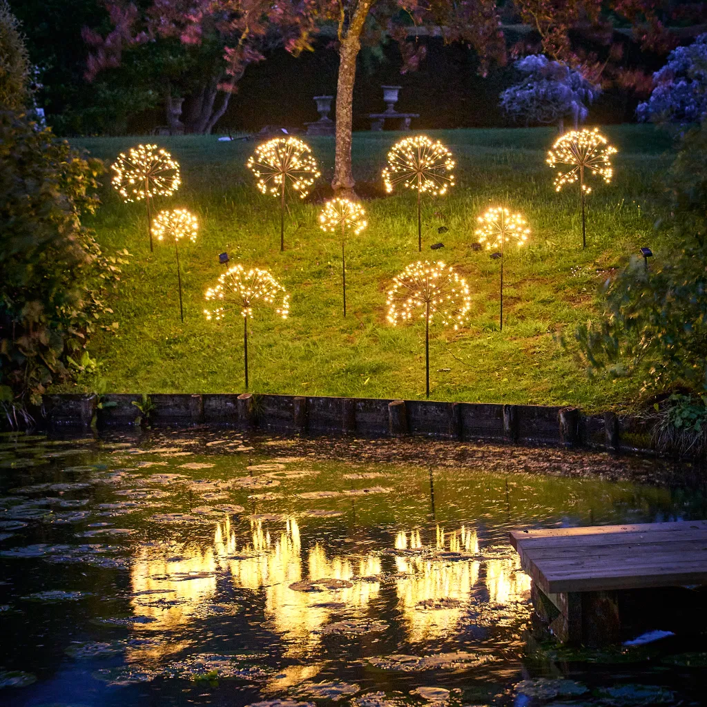 Multiple dandelion-shaped solar stake lights glowing with warm LED light in a lush green garden at dusk. The lights are reflected in a dark pond in the foreground, creating a magical, decorative lighting effect.