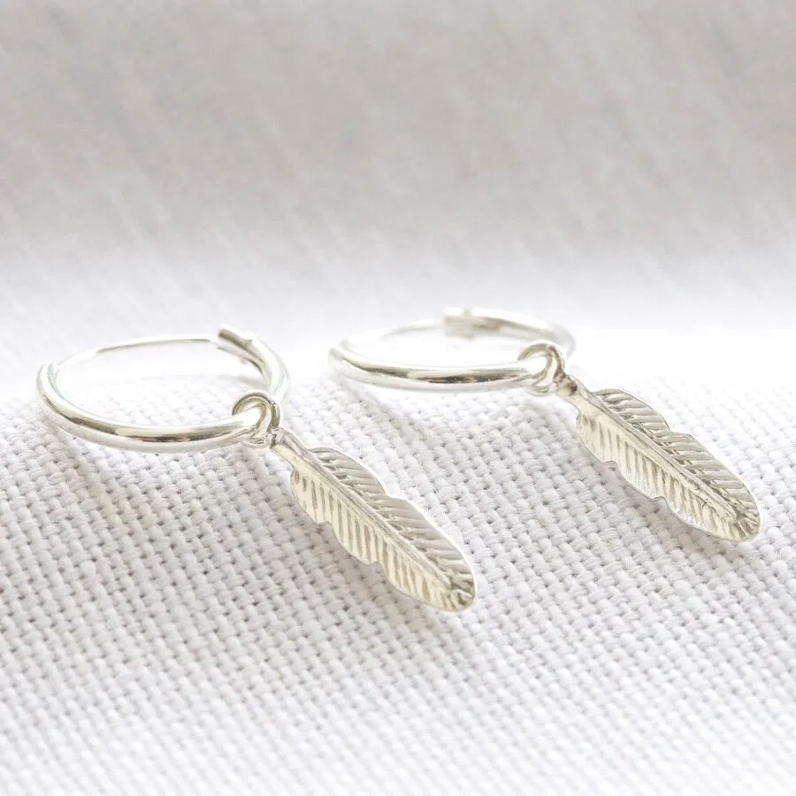 A pair of small, polished sterling silver hoop earrings, each adorned with a delicate, intricately detailed sterling silver feather charm dangling from the base, displayed against a soft, neutral-colored fabric background.