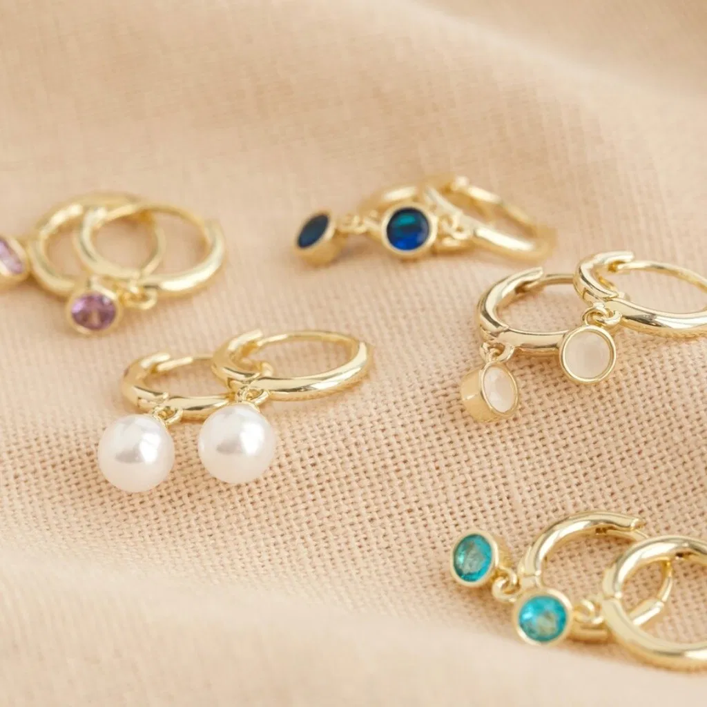 A collection of small gold huggie hoop earrings, each featuring a dangling birthstone or faux pearl charm in various colours (including purple, white, blue, and turquoise), displayed on a neutral textured fabric.