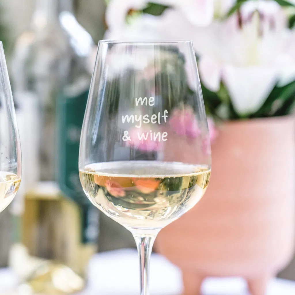 A large, elegant stemmed wine glass filled with white or rosé wine, engraved with the witty slogan 'me myself & wine' in a script font, set outdoors among flowers and a blurred backdrop, ideal for al fresco dining.