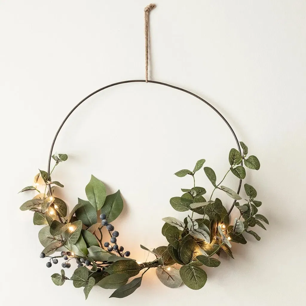Modern Christmas half wreath bundle featuring artificial eucalyptus and dark blue berries, wrapped around a thin black metal hoop and illuminated by warm micro fairy lights, hanging on a white wall by a piece of twine.