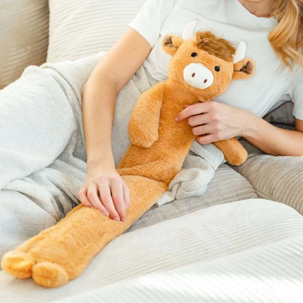 Woman holding a long, cuddly Highland Cow hot water bottle on a sofa, providing warmth and comfort.