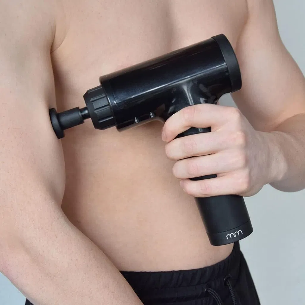Close-up of a man using a black percussive muscle massager gun (with 'mm' logo) on their upper arm, featuring a compact, handheld design for deep tissue recovery.