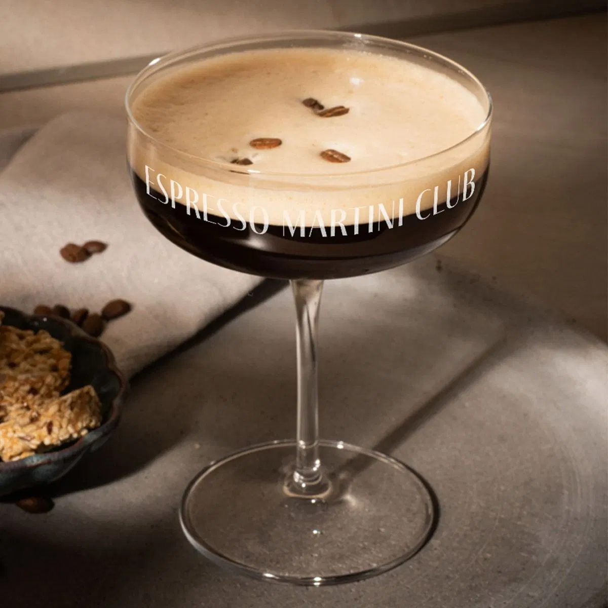 A wide, stemmed coupe glass filled with a dark espresso martini cocktail, topped with a rich layer of foam and three coffee beans, featuring the personalised engraved text 'ESPRESSO MARTINI CLUB' wrapped around the rim.