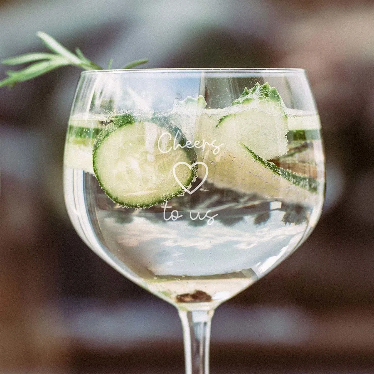 A large, stemmed balloon gin glass (copa) filled with a refreshing gin and tonic, garnished with cucumber slices and a rosemary sprig, featuring the personalised engraved message 'Cheers to us' with a heart symbol in script font.