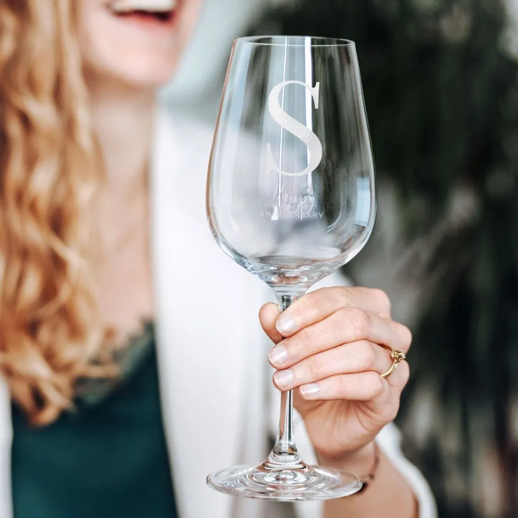 Close-up of a hand holding a clear, elegant wine glass engraved with a large monogram letter 'S' and the text 'HAPPY ANNIVERSARY', held by a woman in a white blazer.