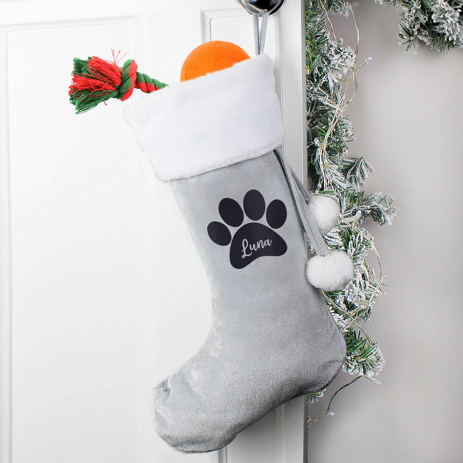 A personalised silver/grey velvet Christmas stocking with a white plush cuff, featuring a large black paw print containing the name 'Luna' in script, hanging from a white door handle and stuffed with a bright orange ball and a red and green rope toy.