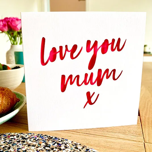 Alt Text: A premium white Mother's Day card featuring "love you mum x" in an elegant, laser-cut cursive script that reveals a vibrant, shimmering pink background underneath, displayed on a wooden table.