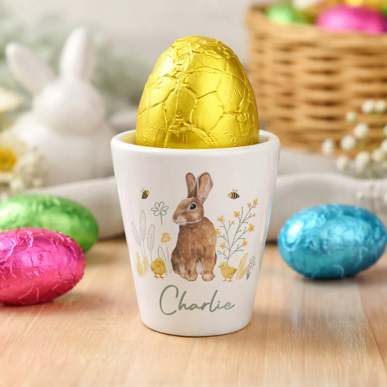 A white ceramic egg cup featuring a charming watercolour illustration of a brown rabbit surrounded by yellow chicks, spring flowers, and bees. The name "Charlie" is printed in an elegant green script at the base. A gold-foil wrapped chocolate Easter egg sits inside the cup, set against a festive background with colourful Easter eggs and a brown bunny figurine.