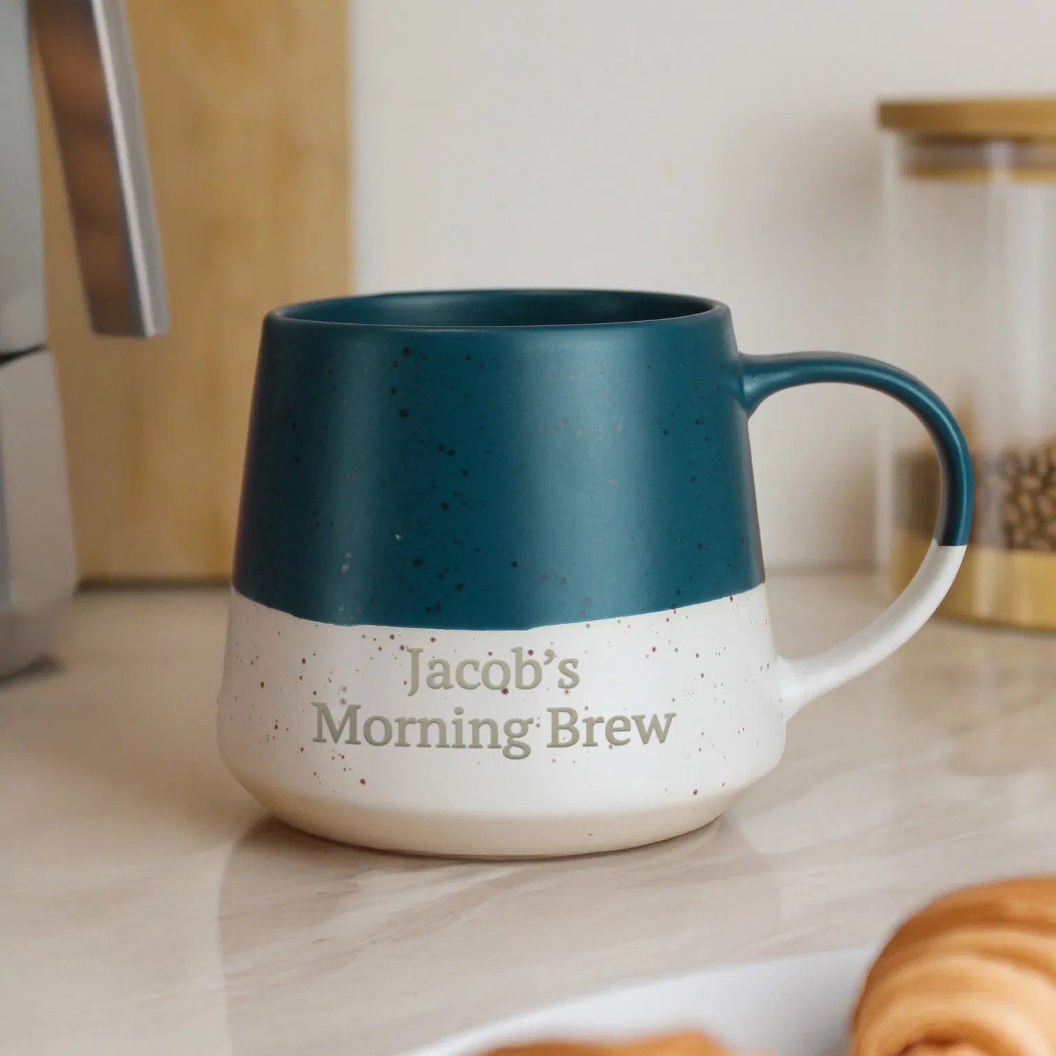A two-toned, tapered ceramic mug with a speckled finish. The top half is a deep matte teal blue, and the bottom half is cream-colored. The cream section is personalized with the text "Jacob's Morning Brew" in a serif font. The mug sits on a marble countertop next to a croissant and a moka pot.