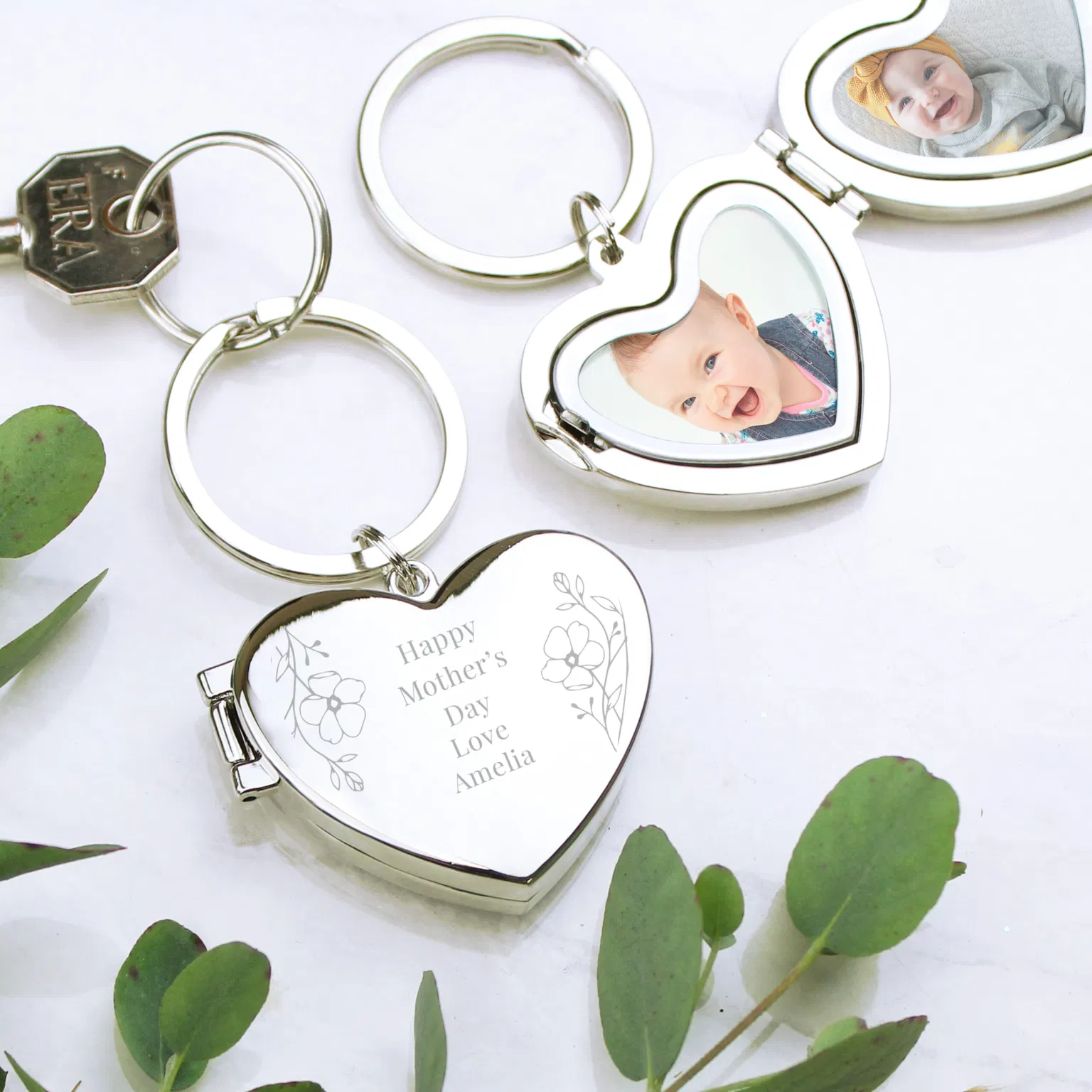 A silver heart-shaped locket keyring shown in three states: closed, partially open, and fully open. The closed locket features an engraved floral design with the text "Happy Motherâs Day Love Amelia." When opened, the locket reveals two heart-shaped photo inserts featuring high-quality portraits of smiling babies. The keyrings are displayed on a white marble surface accented with green eucalyptus leaves.