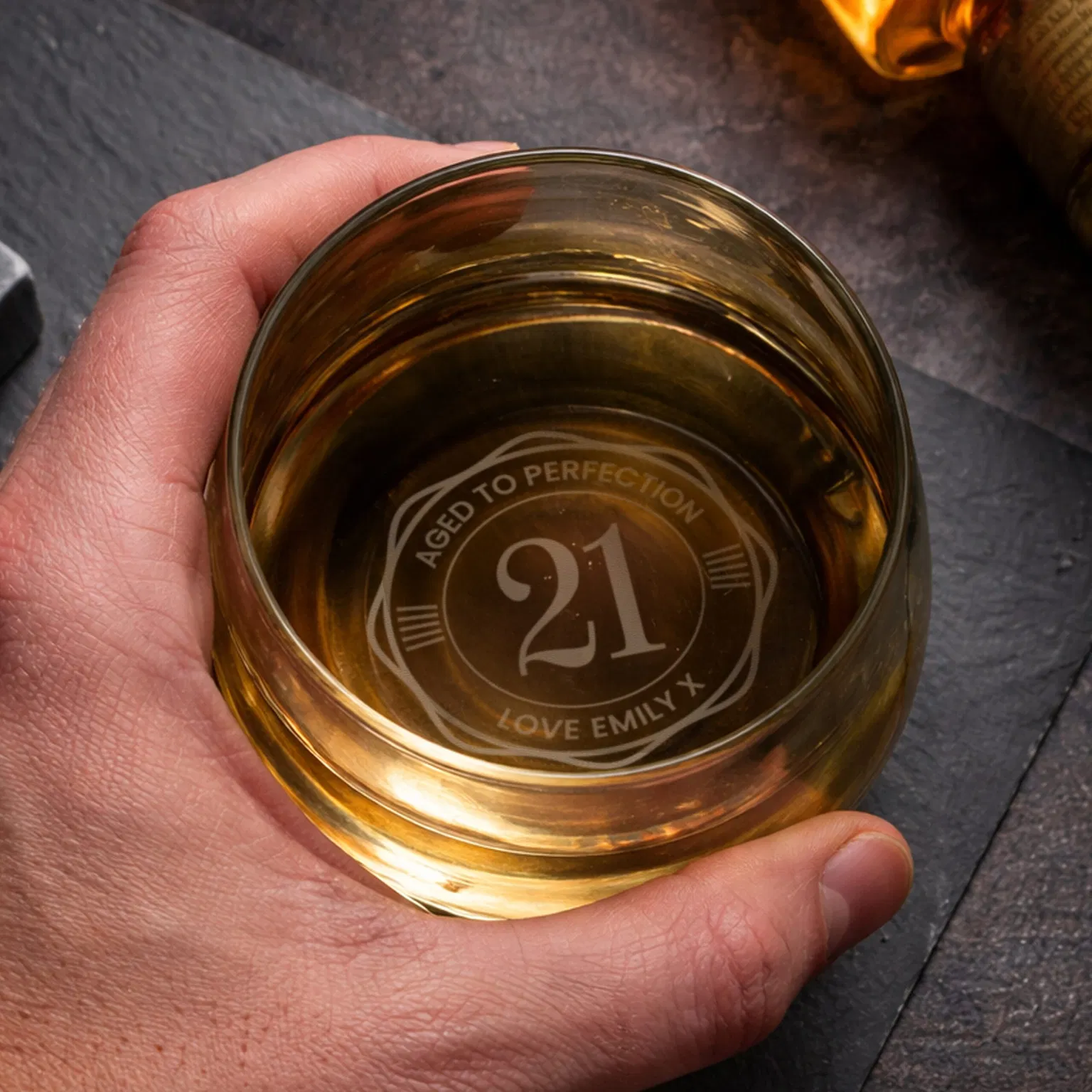 Option 1 (Descriptive): “Hand holding a glass whiskey tumbler filled with amber spirit, showing a personalized circular engraving on the base that reads 'Aged to Perfection 21' with a custom message.