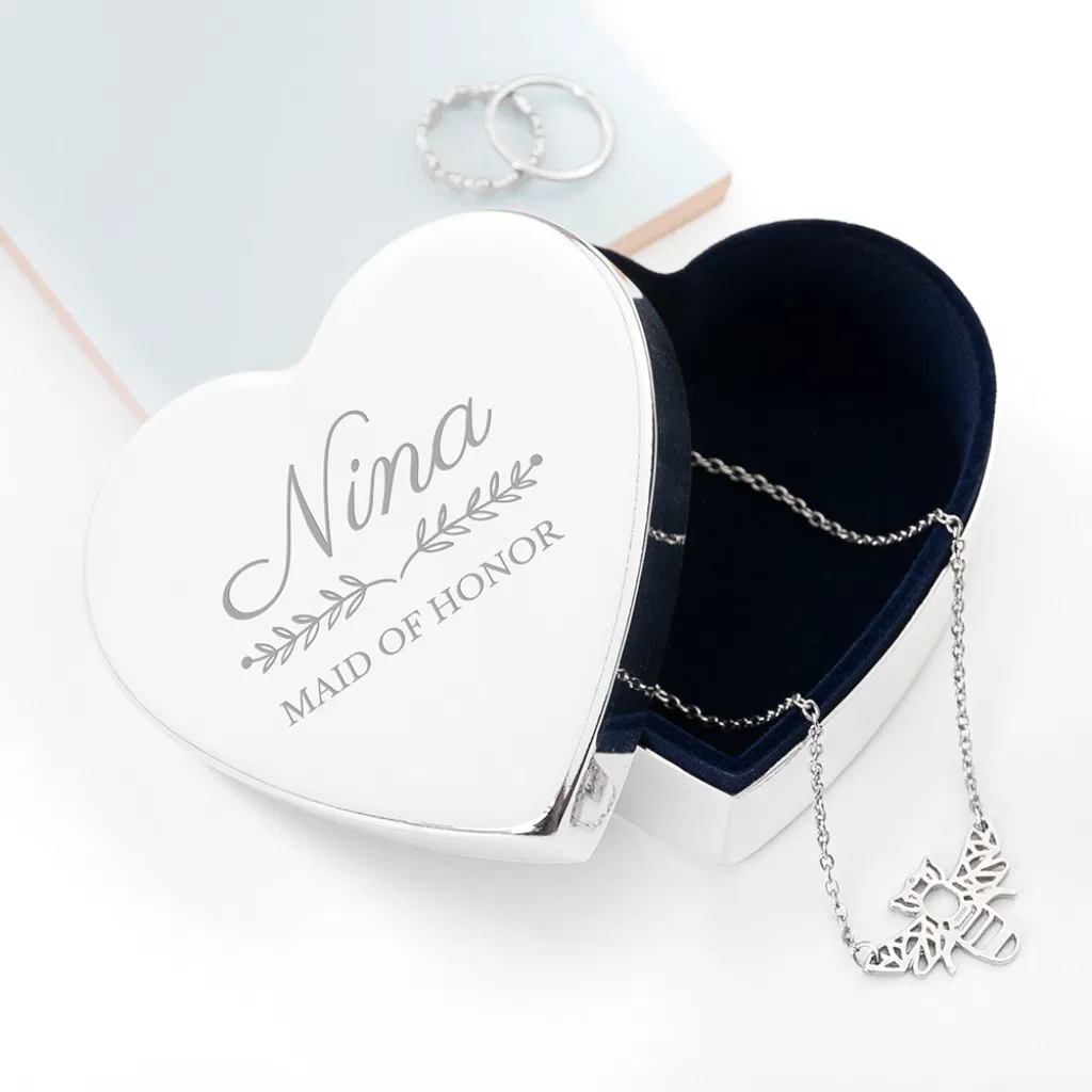 Top-down view of a heart-shaped silver jewellery box with a navy velvet lining. The lid is engraved with "Nina" in a script font and "Maid of Honor" in a serif font, separated by a delicate floral leaf design. A silver bee necklace is draped inside.