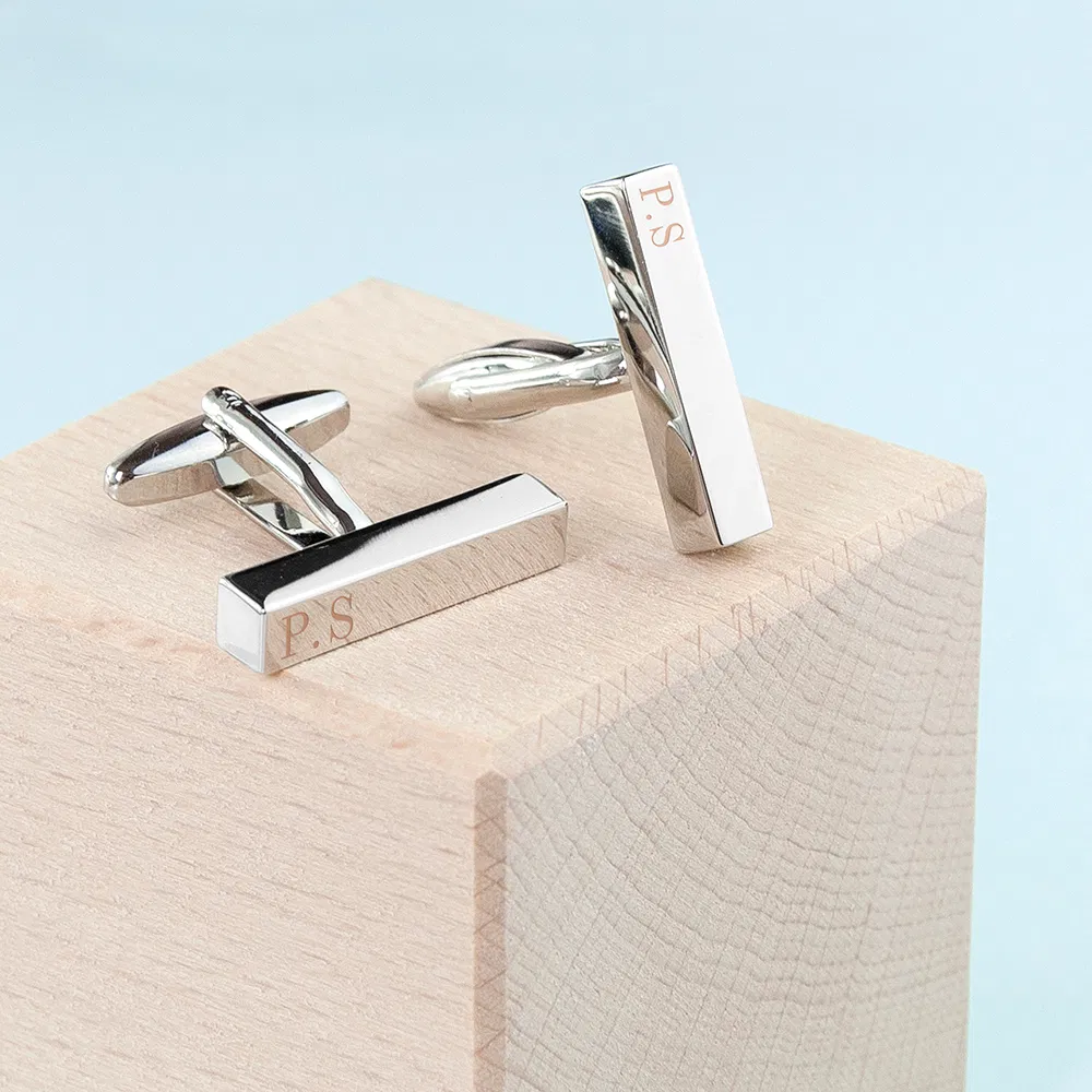 Pair of polished silver bar-shaped cuffhttps://www.google.com/search?q=links with a modern architectural design, featuring custom engraved initials P.S, displayed on a natural light wood block.