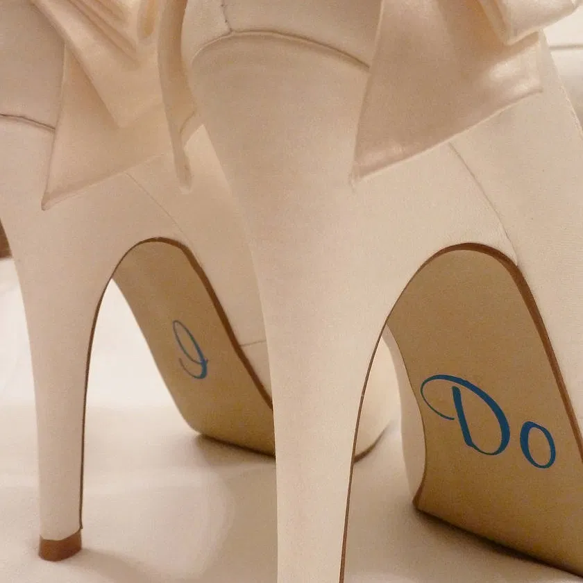 Close-up of ivory high-heeled wedding shoes with silver vinyl stickers on the arches reading "I Do"